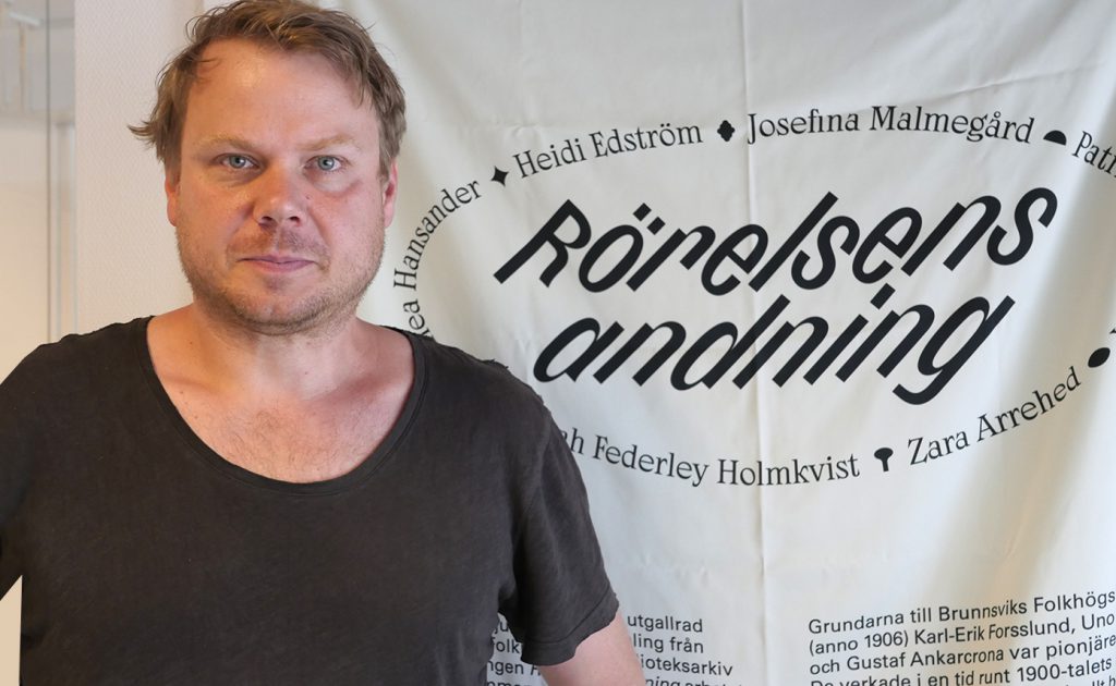 Konstnären Patrick Kretschek i svart t-shirt framför ett sidentyg där utställnningens namn, Rörelsens andning, är tryckt i svart.